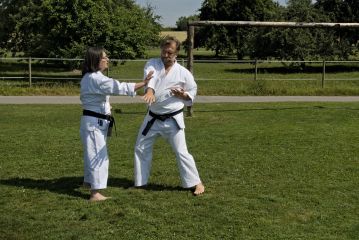 Zwei Karateka üben auf einer Wiese paarweise. Die Sonne scheint.