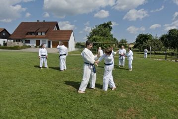 Mehrere Karateka üben auf einer Wiese paarweise. Die Sonne scheint.