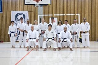 Zehn Karateka stehen in einer Turnhalle aufgereiht, vor ihnen sitzen drei weitere Karateka auf Stühlen.