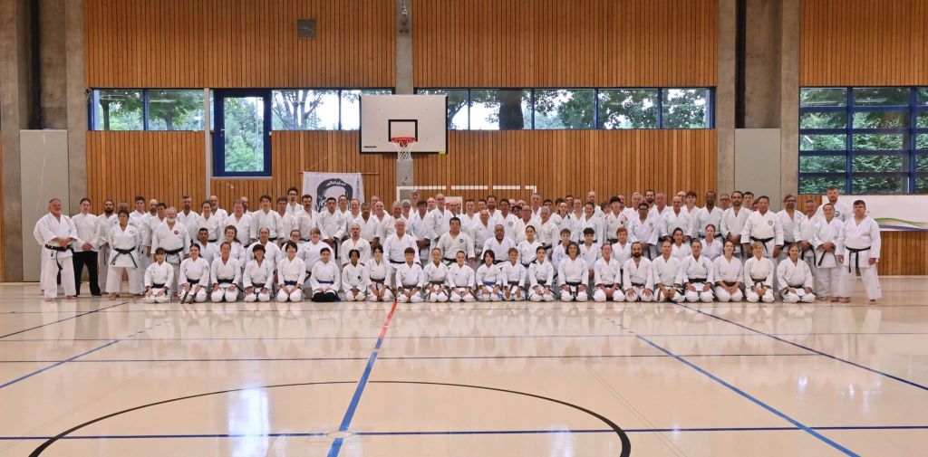 Eine große Gruppe Karateka stehen in einer Turnhalle in mehreren Reihen aufgereiht. Die erste Reihe kniet.