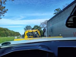 Blick vom Fahrersitz durch die Windschutzscheibe eines Autos. Das Auto wird gerade auf einen gelben Abschleppwagen hochgezogen. Rechts steht ein LKW, links ist die angrenzende Autobahn zu sehen. Zwei Frauen stehen neben dem Abschleppwagen und schauen zu dem Auto hoch.
