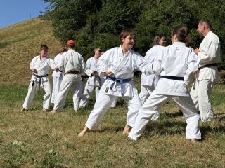 Auf einer Wiese unterhalb eines Hangs stehen Karateka paarweise beieinander. Sie haben jeweils am Unterarm Kontakt zueinander und bewegen ihre Schwerpunkte synchron und abwechselnd vor und zurück.