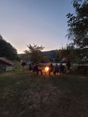 Eine Handvoll Personen sitzen auf Bänken rund um eine Feuerschale, in der ein großes Feuer brennt. Es dämmert. Im Hintergrund sind Bauernhof-Gebäude zu sehen, außerdem ein Blick in ein Tal zwischen steilen, teils bewaldeten Hängen.