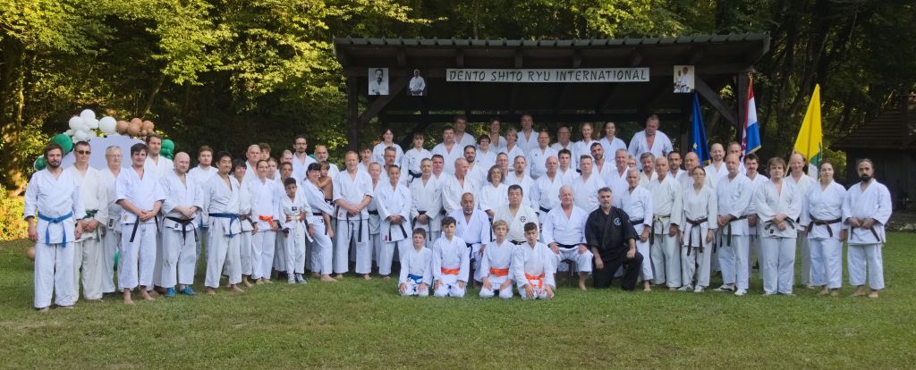 Viele Karateka sind in Reihen auf einer Wiese vor einem Pavillon aufgereiht. Die vordere Reihe kniet im seiza, die hinteren Reihen stehen.