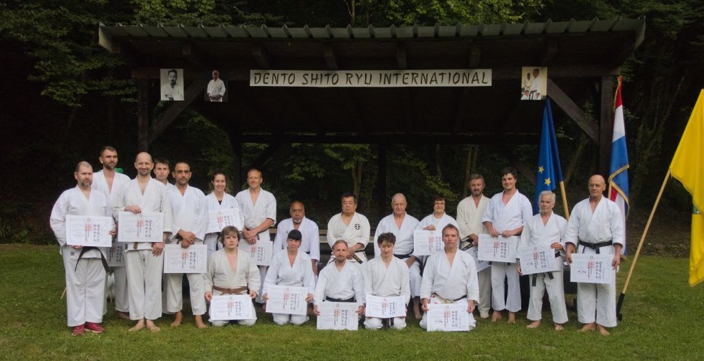 Zwanzig Karateka sind in zwei Reihen vor einem Pavillon auf einer Wiese aufgereiht. Die vordere Reihe kniet im seiza, die hintere Reihe steht. Ein Großteil hält eine große Urkunde mit japanischen Schriftzeichen hoch.